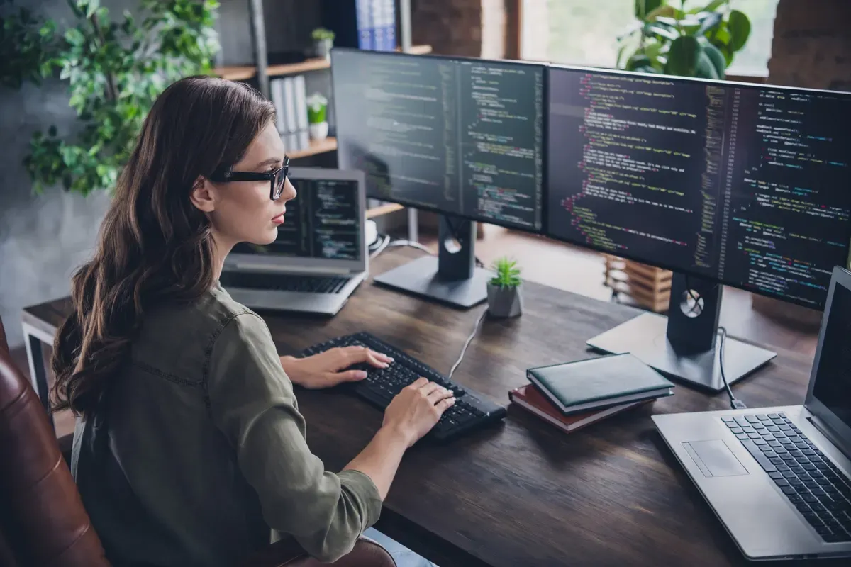 Female developers from unlimited support recovers your site after a crash. She is working at her home office at her desk with two computer screens and two laptops