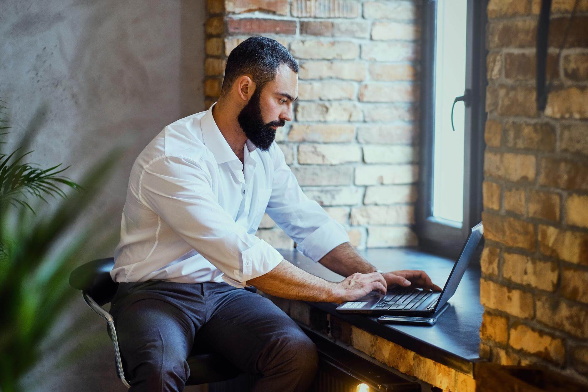 Stylish bearded male works with a laptop in a room with loft interior. Man is writing a report about 5-minute SEO & speed audit for WordPress